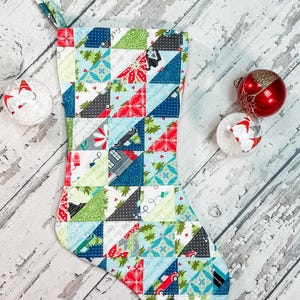 May include: A handmade Christmas stocking with a patchwork design featuring red, green, blue, and gray triangles. The stocking has a loop for hanging and is decorated with Christmas-themed patterns. Two decorative ornaments are next to the stocking.