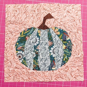 Puede incluir: Un bloque de tela cuadrado para acolchar que presenta un diseño central de calabaza compuesto por varias telas con patrones florales y botánicos en tonos verde azulado, blanco y marrón, sobre un fondo rosa claro con estampado floral.