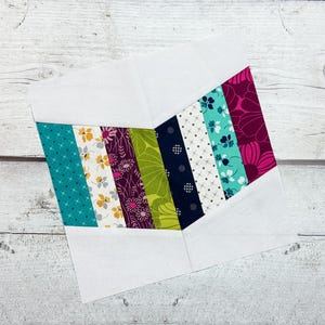 May include: A colorful quilt block featuring a chevron design. The block is composed of various patterned fabric strips in teal, yellow, purple, green, navy, and pink, set against white fabric. The fabrics have floral and geometric patterns.