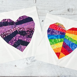 May include: Two heart-shaped quilt blocks. One heart is made of purple and dark blue patterned fabric, the other is a rainbow of colors. Both are set on white fabric with a geometric design.