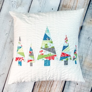 May include: White square pillow with a quilted pattern. The pillow features five colorful patchwork Christmas trees. The trees are made of various patterned fabrics in shades of blue, green, red, and gray. The pillow is on a wooden surface.