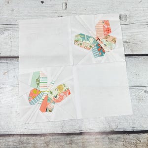 May include: Four white fabric quilt blocks arranged in a square. Each block features a colourful, geometric design in the centre, with floral and patterned fabrics in shades of pink, blue, and green. The blocks are set against a light wood grain background.