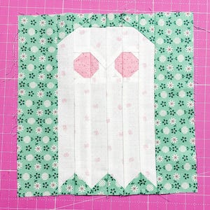 May include: A square fabric quilt block featuring a central white ghost shape with two pink octagonal eyes, constructed from white fabric with a subtle pink floral print, flanked by vertical panels of mint green fabric with a white floral pattern.