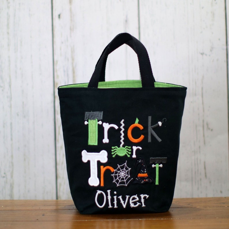 May include: Black trick or treat bag with green, orange, and white embroidered letters and a spider, witch hat, and bone design. The bag has the name "Oliver" embroidered on the bottom.