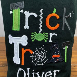 May include: A black canvas trick or treat bag with green, orange, and white embroidered letters spelling "Trick or Treat" and a spider, web, witch hat, and bone. The name "Oliver" is embroidered in white at the bottom.