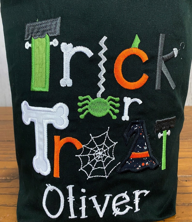 May include: A black canvas trick or treat bag with green, orange, and white embroidered letters spelling "Trick or Treat" and a spider, web, witch hat, and bone. The name "Oliver" is embroidered in white at the bottom.