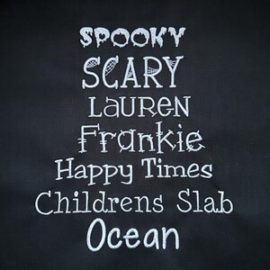 May include: A black fabric tote bag with white embroidered text that reads "Spooky Scary Lauren Frankie Happy Times Childrens Slab Ocean".