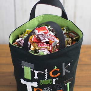 May include: Black trick or treat bag with green and orange embroidery. The bag is filled with various candy bars, including Twix, Krackel, and Snickers. The bag is personalized with the name "Oliver".