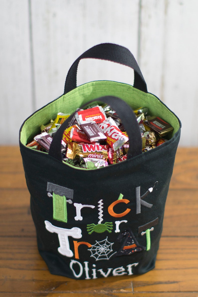 May include: Black trick or treat bag with green and orange embroidery. The bag is filled with various candy bars, including Twix, Krackel, and Snickers. The bag is personalized with the name "Oliver".