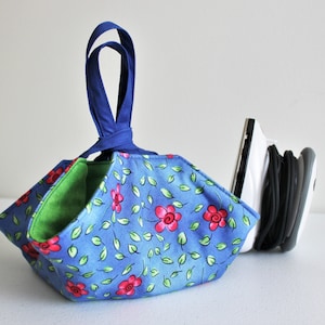 May include: A blue and green fabric storage basket with a blue handle. The basket is folded into a triangular shape and has a floral pattern with red flowers and green leaves. The basket is sitting on a white surface.