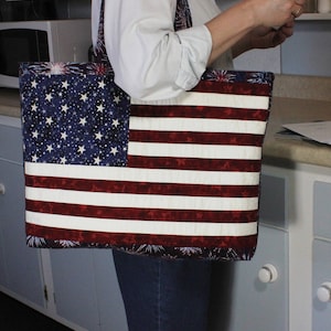 Puede incluir: Una bolsa de mano roja, blanca y azul con un diseño de estrellas y rayas. La bolsa tiene un fondo azul con estrellas blancas y rayas rojas y blancas. La bolsa está hecha de tela y tiene una cremallera.