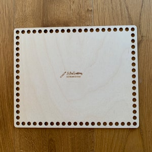 Könnte beinhalten: Ein heller Holzwebrahmen mit rechteckiger Form und abgerundeter Kante. Der Webrahmen hat Löcher um den Umfang herum zum Weben von Garn. Der Webrahmen trägt den Text "Le Loup Creations" und "DIY MAKE YOUR OWN INCHCRAFT".