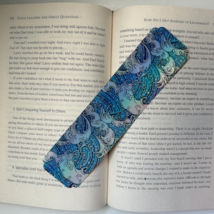 May include: A blue and white bookmark with a wave pattern. The bookmark has a holographic finish and is decorated with white stars. The bookmark is on top of an open book.