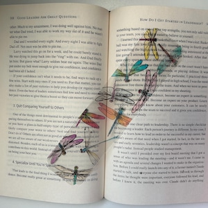 May include: An open book with a page turned down. The page is decorated with colorful dragonfly illustrations. The text on the page reads "Good Leaders Ask Great Questions" and "How Do I Get Started in Leadership?"