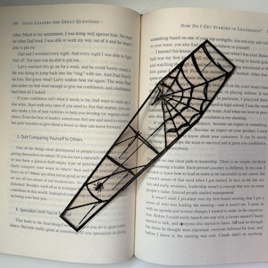 May include: A black metal bookmark shaped like a spider web with a spider in the center. The bookmark is placed on an open book with text about leadership.
