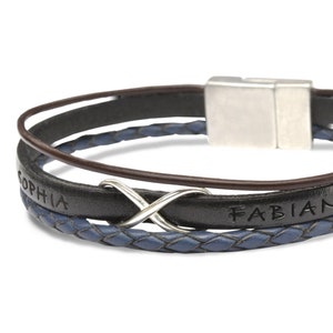 Könnte beinhalten: Ein mehrsträngiges Armband mit einem glatten schwarzen Lederband, in das die Namen Sophia und Fabian eingraviert sind, einer geflochtenen blauen Lederschnur und einem dünnen braunen Lederband. Es enthält ein silbernes Unendlichkeitssymbol und einen silbernen Magnetverschluss.