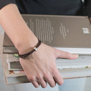 May include: A brown leather bracelet with a silver charm. The bracelet is worn on a person's wrist.