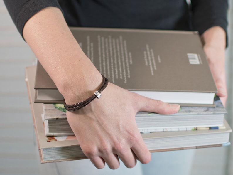 May include: A brown leather bracelet with a silver charm. The bracelet is worn on a person's wrist.