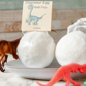 May include: Two white bath fizzies shaped like dinosaur eggs on a white plate with a dinosaur toy and a label that reads "Dinosaur Egg Bath Fizzy".