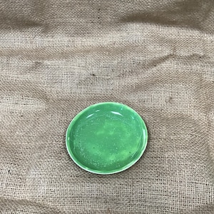 May include: A small, round, green ceramic dish with an irregular shape. The dish has a glossy finish and a white rim. The background is a rough, tan burlap fabric.