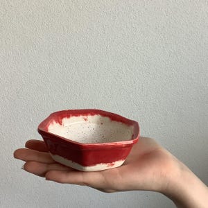 May include: A small, handmade ceramic bowl with a distinctive shape. The bowl features a bright red exterior and a speckled white interior. The rim has a textured, uneven edge. The bowl is held in a person's hand.