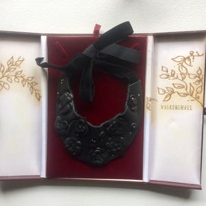 May include: A black leather collar necklace with embossed floral details, presented in an open box with a red velvet interior. The box has gold leaf accents and the text "WALKERHALL". A black ribbon is attached to the necklace.