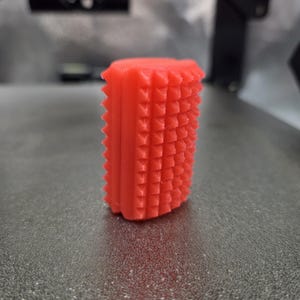 Puede incluir: Un objeto rectangular rojo brillante con una superficie texturizada. El objeto tiene pequeñas protuberancias en forma de pirámide en sus lados. Los bordes superior e inferior tienen un diseño dentado. El objeto parece estar impreso en 3D.