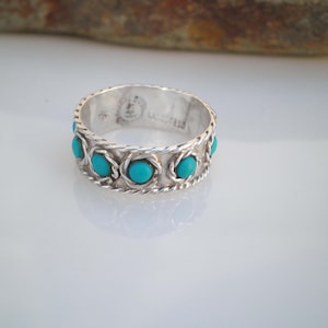 Puede incluir: Un anillo de plata con una banda trenzada y piedras turquesa engastadas en la banda. El anillo está marcado con "STERLING".