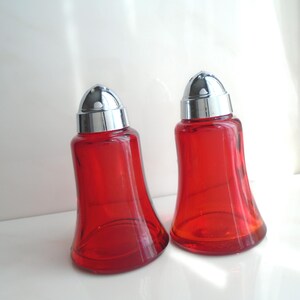 Könnte beinhalten: Zwei rote Salz- und Pfefferstreuer aus Glas mit verchromten Deckeln. Die Streuer haben ein glockenförmiges Design und stehen auf einer weißen Oberfläche. Die Streuer sind leer.