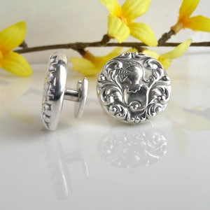 May include: A pair of silver cufflinks with an ornate, engraved design. One cufflink is shown on its side, revealing a textured edge. The other cufflink displays a detailed relief of a figure within a swirling pattern. Yellow flowers are in the background.