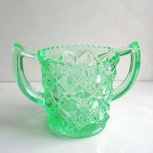 May include: A vintage, light green glass sugar bowl with two handles. The bowl has a diamond pattern and a scalloped rim. The glass is a vibrant, translucent green.