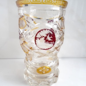 Può includere: Un vaso di vetro trasparente con un design strutturato in vetro tagliato. Il vaso presenta accenti dorati e un emblema circolare rosso e bianco con una scena paesaggistica. Il bordo è anch'esso rifinito in oro. Un pezzo decorativo per l'arredamento della casa.