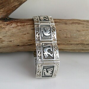 May include: A silver-toned bracelet with rectangular links, each featuring a raised design of birds and other figures. The bracelet is displayed on a piece of driftwood, showcasing its intricate details and craftsmanship.