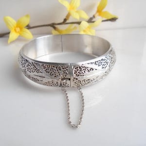 May include: A silver filigree hinged bangle bracelet with intricate openwork patterns. The bracelet has a safety chain and is displayed on a white surface with yellow flowers in the background. The bracelet's design is detailed and elegant.