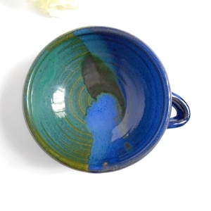 Puede incluir: Un cuenco de sopa de cerámica hecho a mano con un asa en forma de lazo. El cuenco presenta una vibrante paleta de colores de azules, verdes y un toque de amarillo. El interior es una superficie brillante y reflectante.
