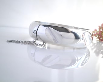 May include: A polished silver-tone hinged bangle bracelet with a smooth, reflective surface and a series of subtle, rounded indentations along the outer band. The bracelet features a secure clasp mechanism and a delicate attached safety chain.