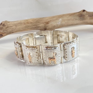 May include: Silver-tone bracelet with rectangular links, featuring gold-tone figures and geometric patterns. The bracelet has a polished finish and is displayed on a reflective surface. The design incorporates religious or historical imagery.