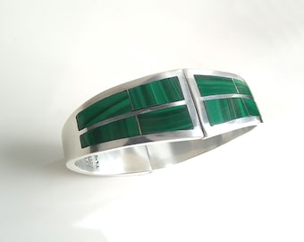 May include: A silver-toned hinged bracelet with rectangular malachite inlays. The bracelet features a polished finish and a secure clasp. The green malachite stones are set in a geometric pattern, creating a striking contrast against the silver.