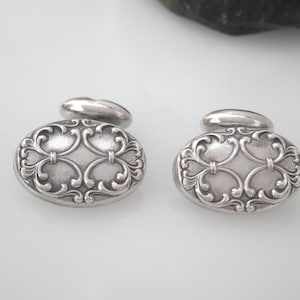 May include: A pair of silver cufflinks with an ornate, oval-shaped design. The cufflinks feature a detailed, intricate pattern of swirls and curves.