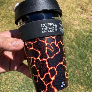 Puede incluir: Una taza de café reutilizable con una tapa negra y un diseño de flujo de lava. La taza tiene una banda blanca con el texto "COFFEE THE WAY IT SHOULD BE".
