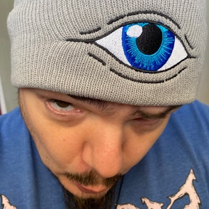 May include: A gray beanie with a blue eye embroidered on the front. The eye has a black pupil and a white sclera.
