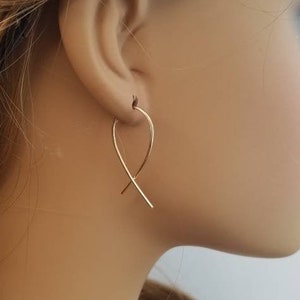 May include: Gold-colored, minimalist earrings in a curved, intersecting design. The earrings are suspended from the earlobe with a simple hook. The image shows the earrings on a person's ear, highlighting their delicate and elegant form. The earrings are a simple, yet stylish accessory.