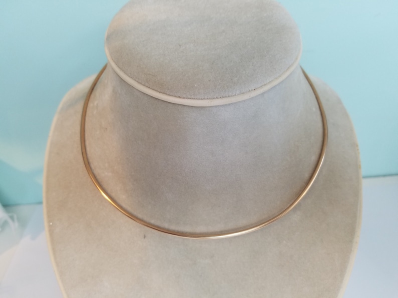 K&ouml;nnte beinhalten: Eine einfache goldene Choker-Kette auf einer wei&szlig;en Mannequin-B&uuml;ste.