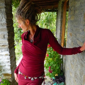 May include: A woman with dreadlocks wears a maroon wrap top with a silver necklace and a silver and amber beaded belt. She is standing in front of a stone wall with greenery in the background.