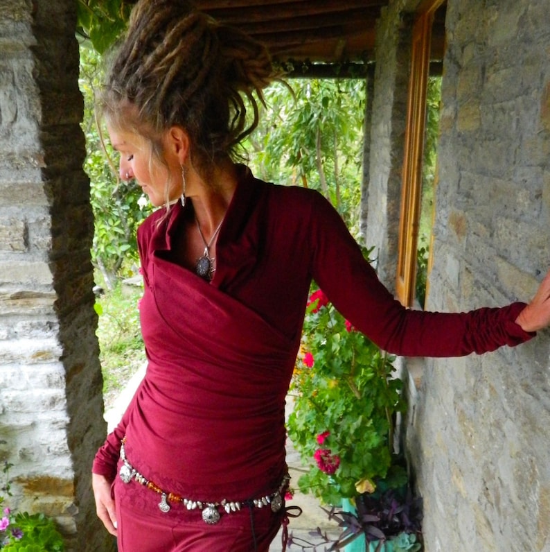 May include: A woman with dreadlocks wears a maroon wrap top with a silver necklace and a silver and amber beaded belt. She is standing in front of a stone wall with greenery in the background.