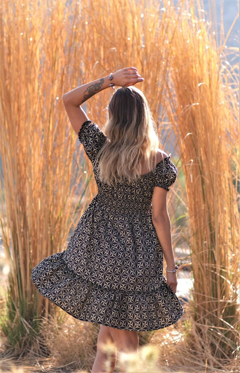 May include: A woman wearing a black and white patterned dress with a ruffled hem. She is standing in front of a field of tall, dry grass.