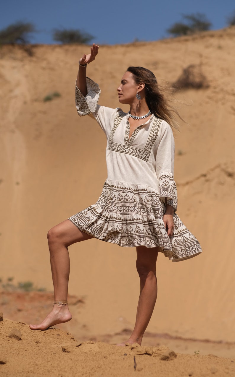 May include: Cream-colored mini dress with long sleeves and a V-neckline. The dress features a geometric pattern in brown around the neckline, sleeves, and hem. The model is standing on a sandy surface.
