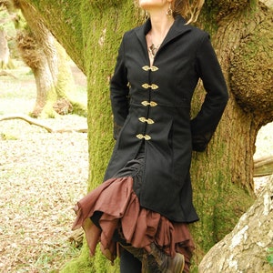 May include: A woman wearing a black coat with gold buttons and a brown ruffled skirt. She is standing in a forest with a large tree behind her.