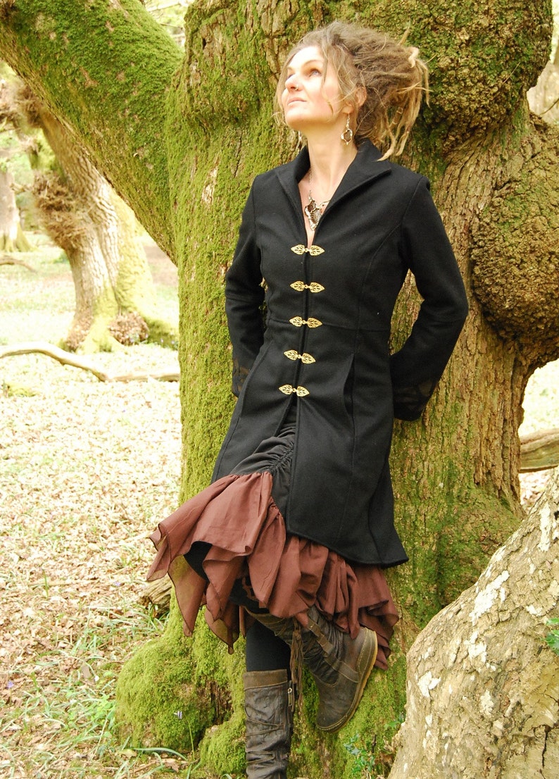 May include: A woman wearing a black coat with gold buttons and a brown ruffled skirt. She is standing in a forest with a large tree behind her.
