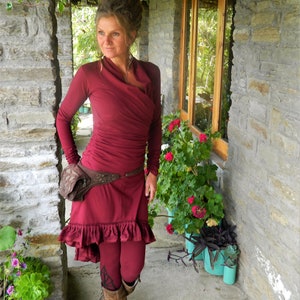 May include: A woman wearing a maroon wrap dress with a brown leather belt and brown boots. She is standing in a doorway with plants and stone walls.